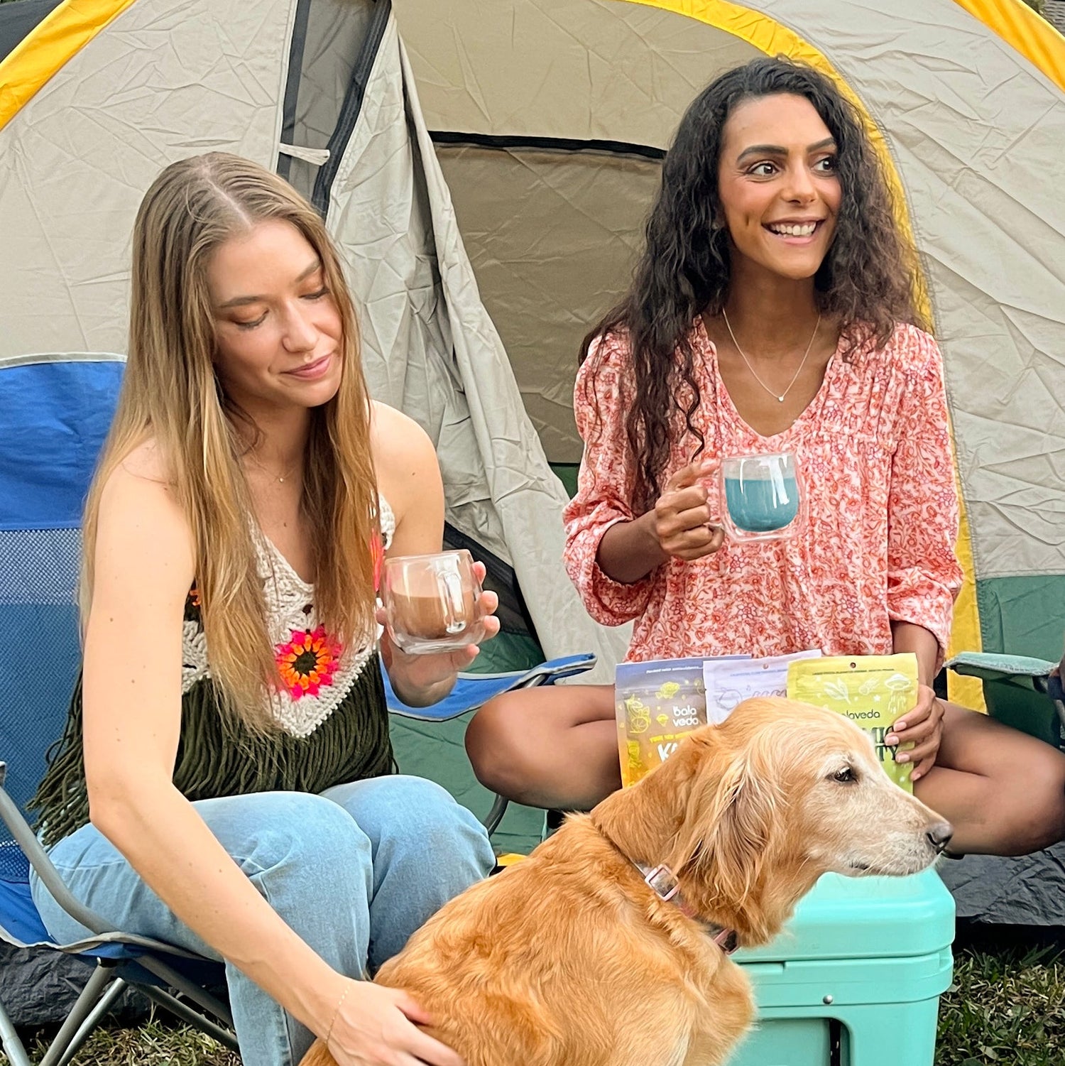 Two women with a dog sitting outside a tent, holding balaveda drinks.