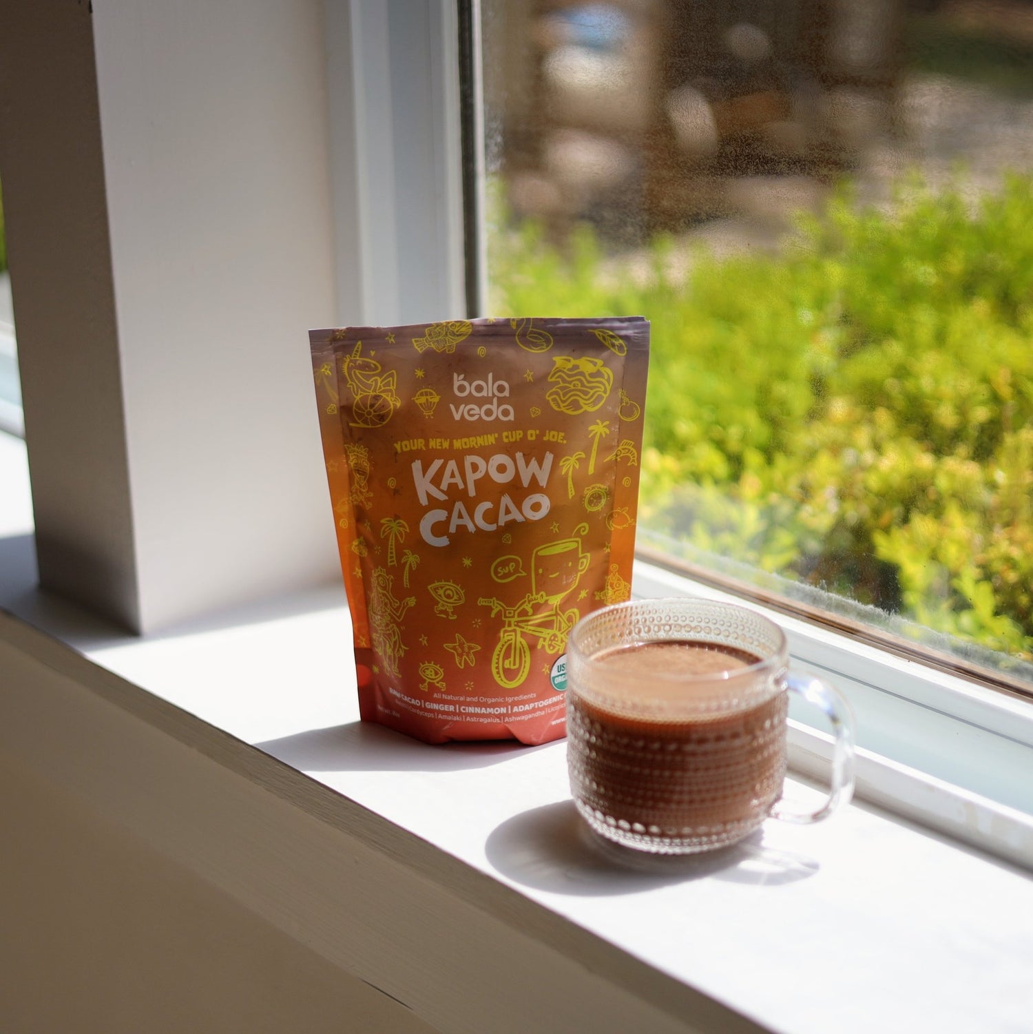 Bala Veda Kapow Cacao package and a glass of cacao on a windowsill with a view of greenery outside.