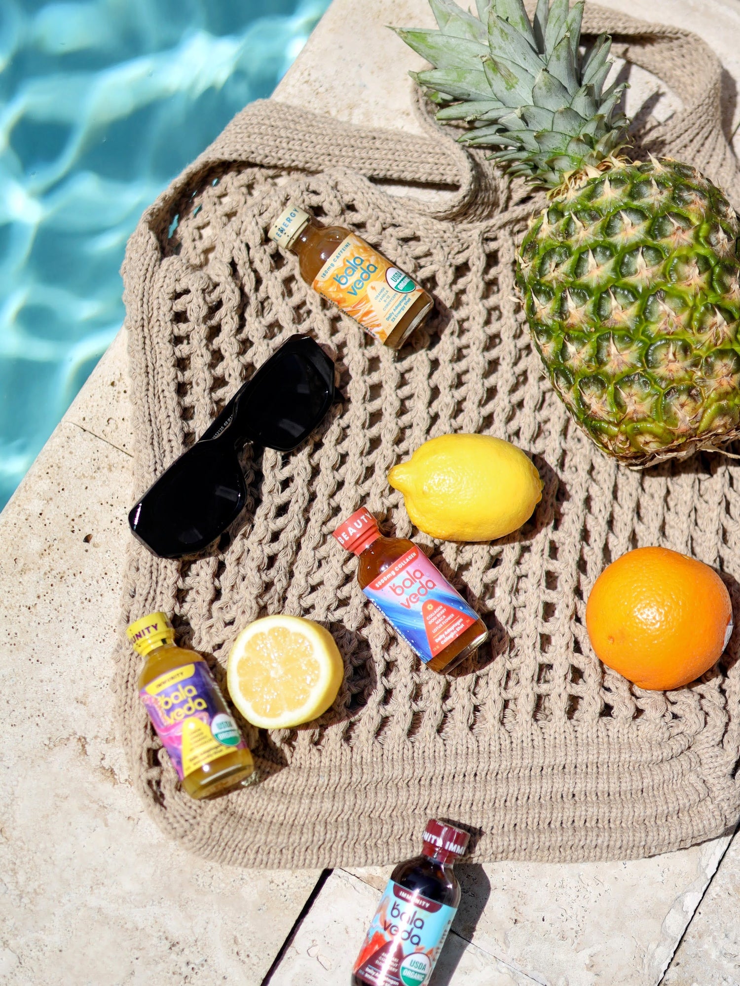 balaveda beauty, immunity, and energy shots sitting on a woven bag with fruit and sunglasses, by a swimming pool