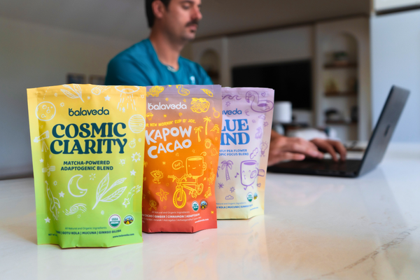 Three Balaveda functional beverage powder packages on a table with a person using a laptop in the background.