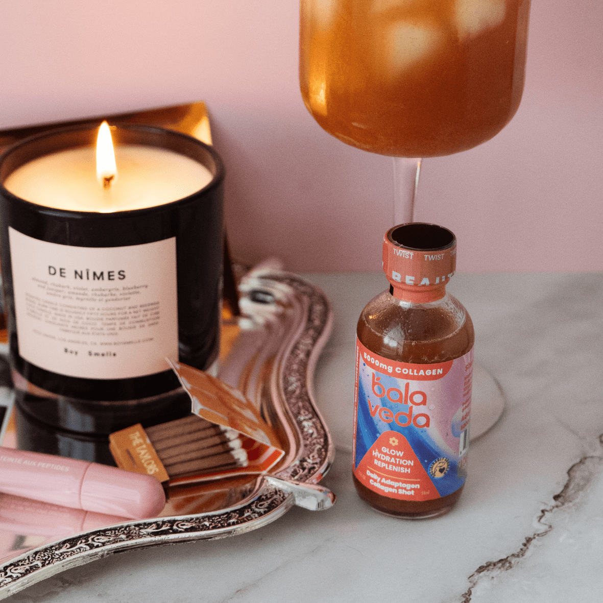 balaveda Radical Beauty Collagen Shot in glass bottle with wine glass behind it and candle and matches on a tray next to it on marble counter.