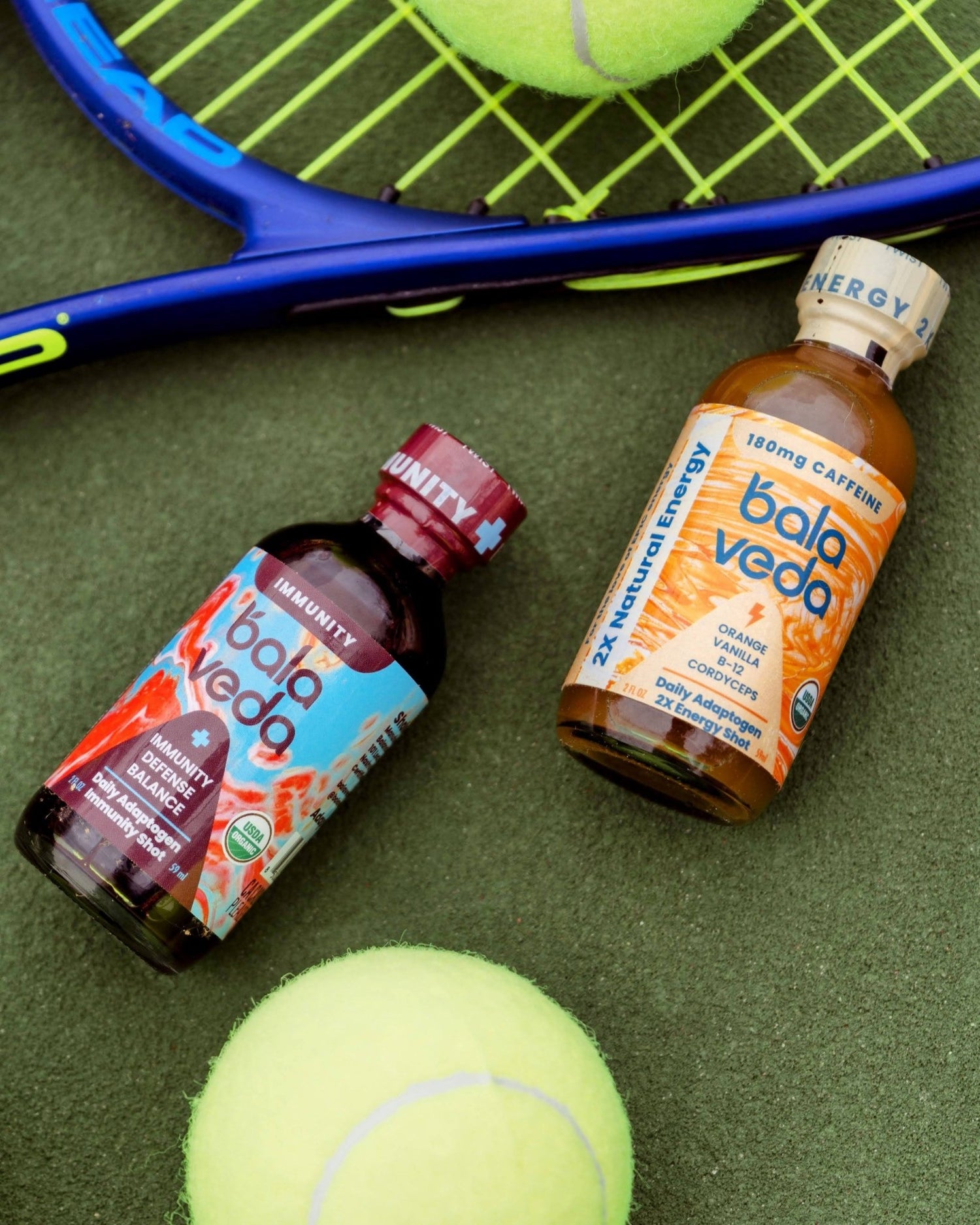 2 balaveda Supersonic Citrus 2x Energy Shot with tennis racket and tennis ball on green background.