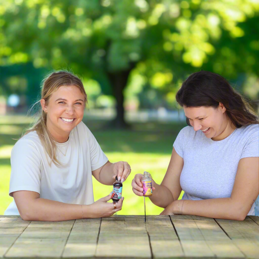 Two women sitting at a wooden table outdoors, smiling and enjoying balaveda immunity shots.
