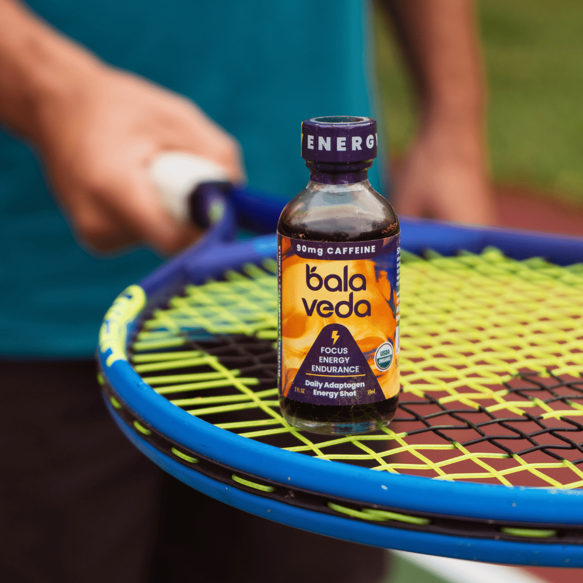 Bottle of Balaveda Wicked Blueberry Energy Shot standing on yellow and blue tennis racket with person in the background holding the handle.