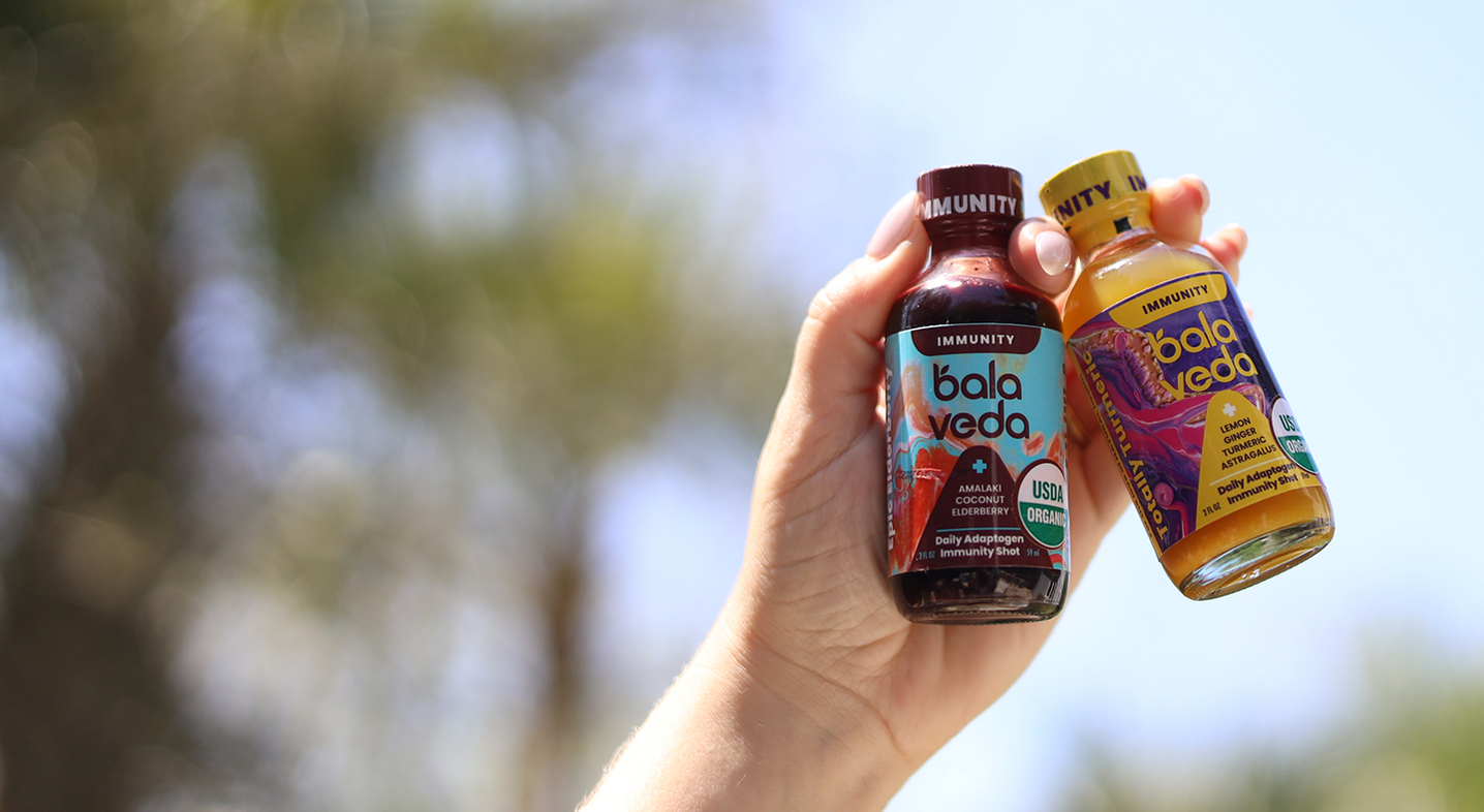 Hand holding two bottles of balaveda immunity shots against a blurred natural background