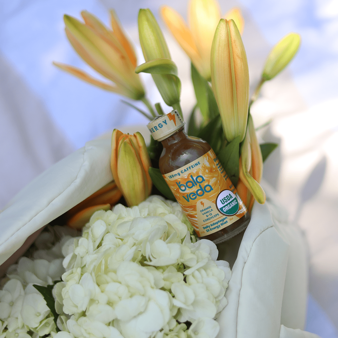 Bala Veda supersonic citrus natural supplement energy shot bottle among flowers with a blurred background