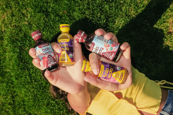 Hands holding multiple bottles of balaveda immunity shots on grass