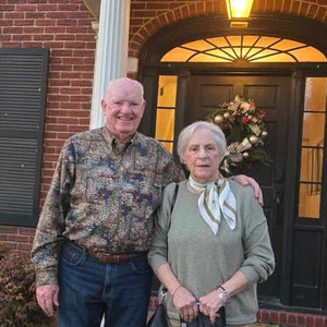 Two elderly people standing in front of a brick building with a decorative wreath on the door.