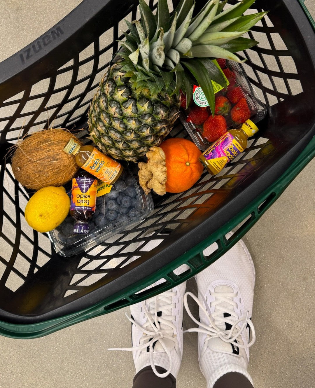 Shopping basket filled with fruits, roots, and balaveda drinks over a concrete floor.