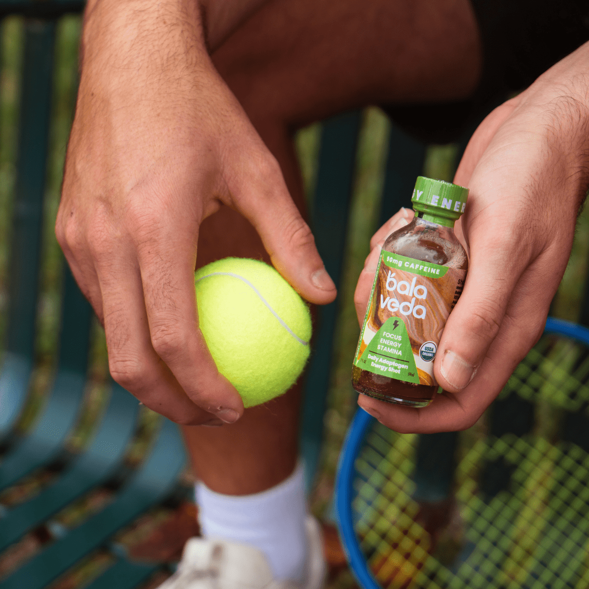 One hand holding a tennis ball and one hand holding a Balaveda Tropic Rush Energy Shot in orange and green bottle. Person sitting on bench with tennis racket in background. 